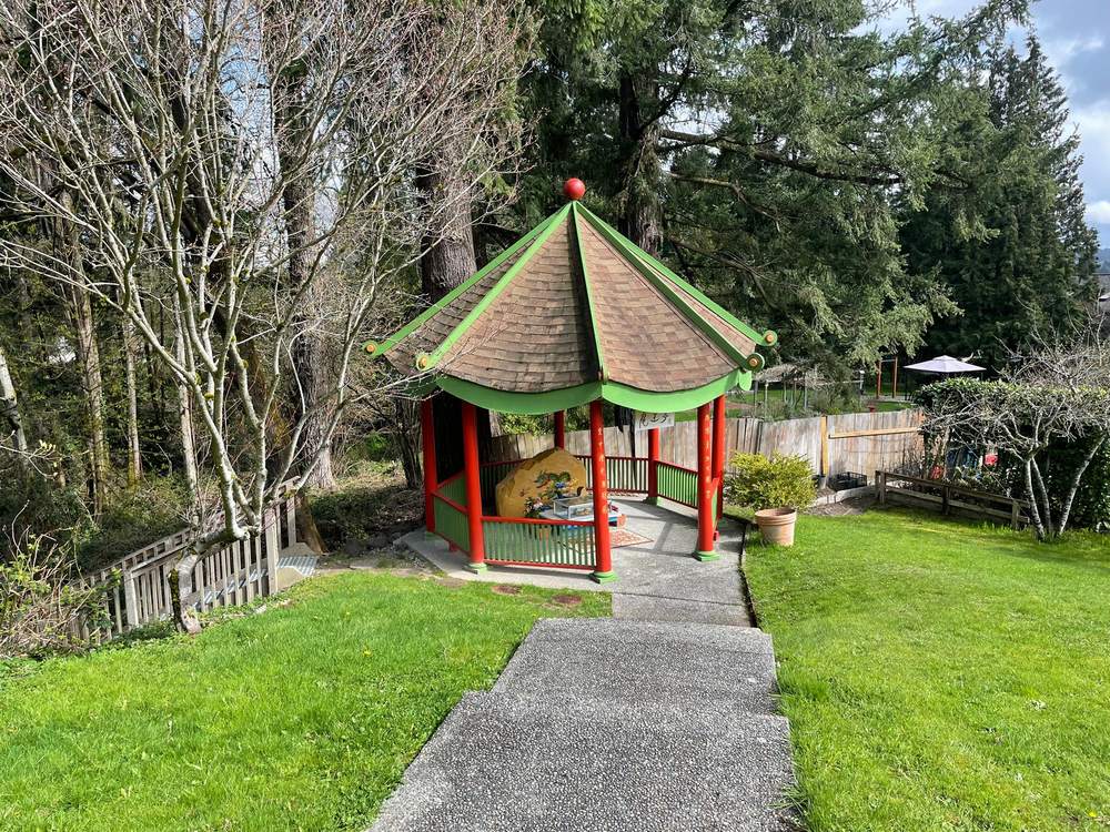 Un petit pavillon ouvert aux colonnes rouges et au toit bordé de vert, sur une pelouse parmi de grands arbres, une pierre dorée visible à l'intérieur