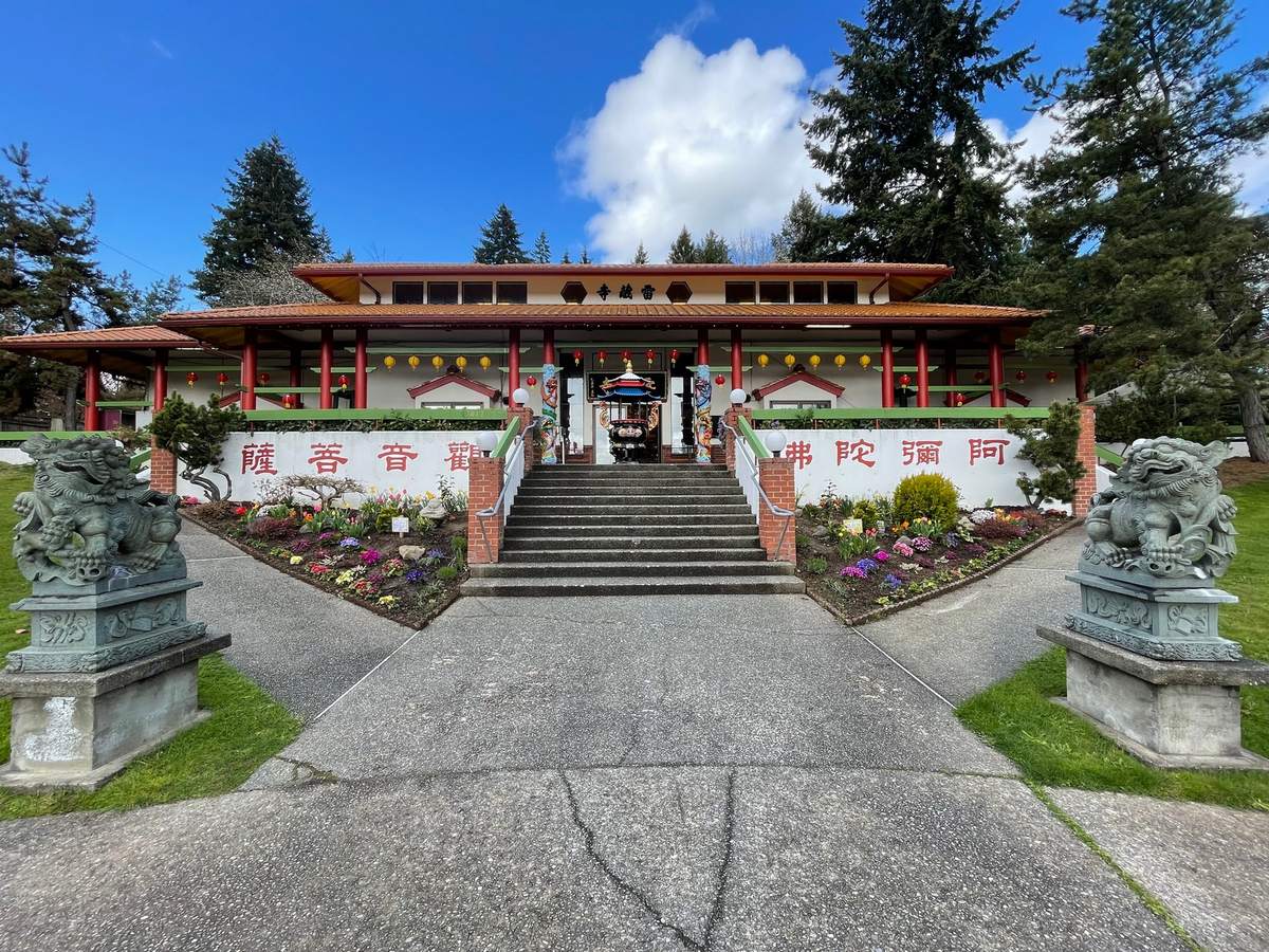La façade du Temple Ling Shen Ching Tze à Redmond : des colonnes rouges sous un toit de tuiles, des parterres de fleurs de part et d'autre de l'escalier et deux lions gardiens de pierre