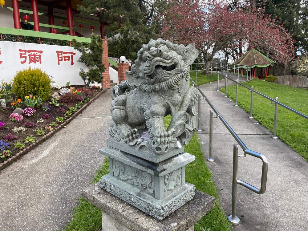 Un lion gardien de pierre sculptée sur un piédestal, au bord du chemin menant à la salle principale, le toit du Pavillon du Roi Dragon visible parmi les arbres à l'arrière-plan